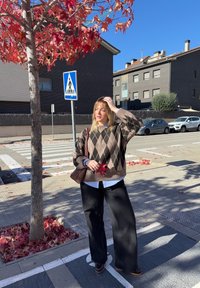 Hnedý sveter s kockovaným vzorom ponad bielu košeľu, skombinovaný s čiernymi širokými nohavicami a hnedými topánkami. Červené listy obklopujú dopravnú značku pre chodcov a strom.