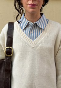 Femme portant un pull crème à col en V sur une chemise à rayures bleues et blanches avec une bandoulière de sac en cuir marron sur l'épaule, devant un mur beige.