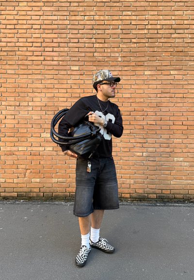 Hombre con gorra negra con estampado, gafas, camiseta deportiva negra, pantalones cortos vaqueros y zapatos con diseño de tablero de ajedrez, sosteniendo una bolsa negra contra una pared de ladrillos.
