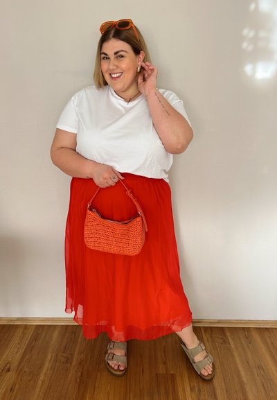 Falda roja en capas, camiseta blanca, bolsa naranja y gafas de sol. El material parece ligero y fluido. Las sandalias son beige con detalle de hebilla.