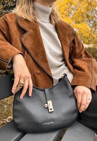 Bolsa de mão em pele negra com uma textura suave, com acabamentos em prata e fecho em zíper. Logótipo da marca "LANCASTER PARIS" apresentado na frente.