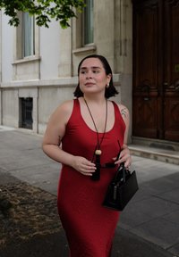 Donna in un vestito rosso senza maniche che tiene occhiali da sole e una borsa nera, in piedi su un marciapiede cittadino vicino a un edificio con porte in legno.
