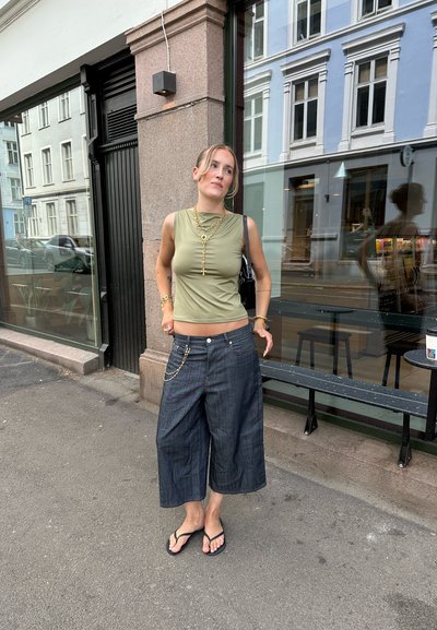 Top sin mangas verde con escote redondo, combinado con culottes de mezclilla oscura sueltos que tienen un acento de cadena; sandalias negras tipo flip-flop.
