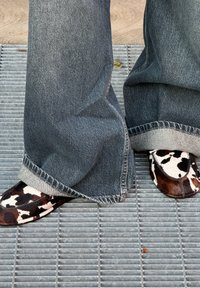 Tehénmintás slip-on cipők lekerekített orral és sötétbarna részletekkel, széles szárú farmernadrággal, amelynek szegélye rojtos.