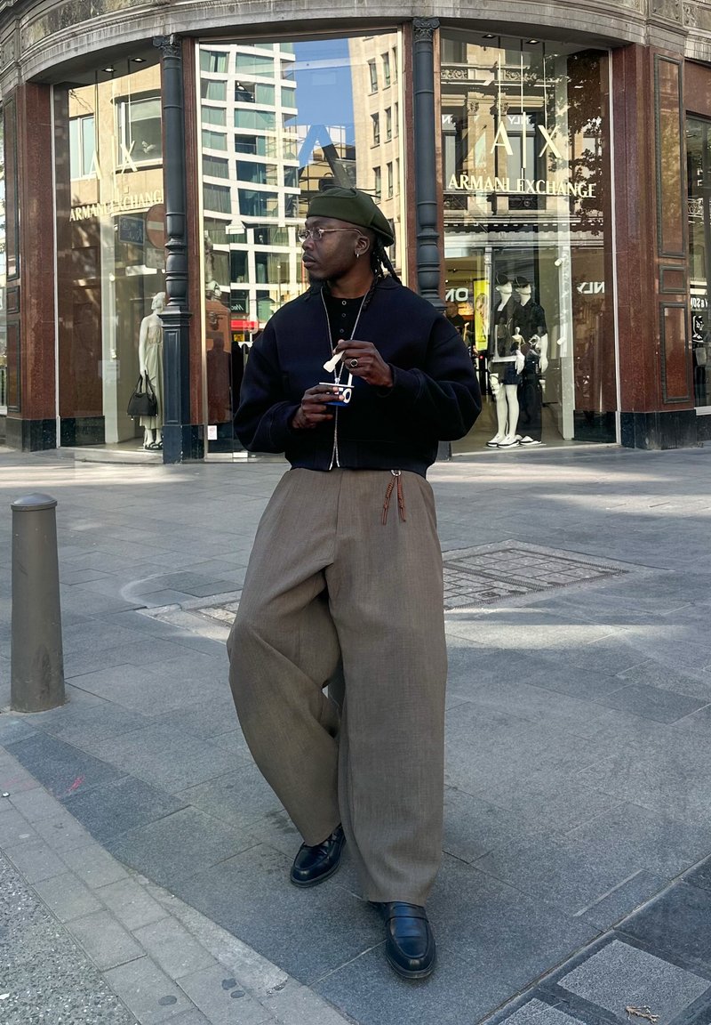 Persona con pantaloni marroni larghi, giacca nera e basco verde, che tiene una macchina fotografica, in piedi davanti al negozio Armani Exchange su una strada cittadina.