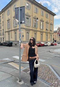 Žena v béžovom voľnom blejzri, čiernom tielku a námorníckych širokých džínsoch drží krémovú tašku a stojí pri dopravnej značke vedľa žltej budovy.