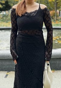 Haut en dentelle noire à manches longues, présentant un motif texturé et des découpes sur les épaules. Porté avec une jupe noire texturée assortie.