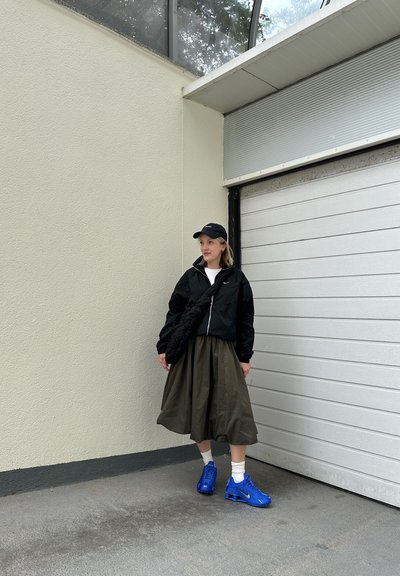 Chaqueta negra con cremallera, camiseta blanca, falda verde olivo, zapatillas azules y gorra negra, de pie contra una puerta de garaje blanca.