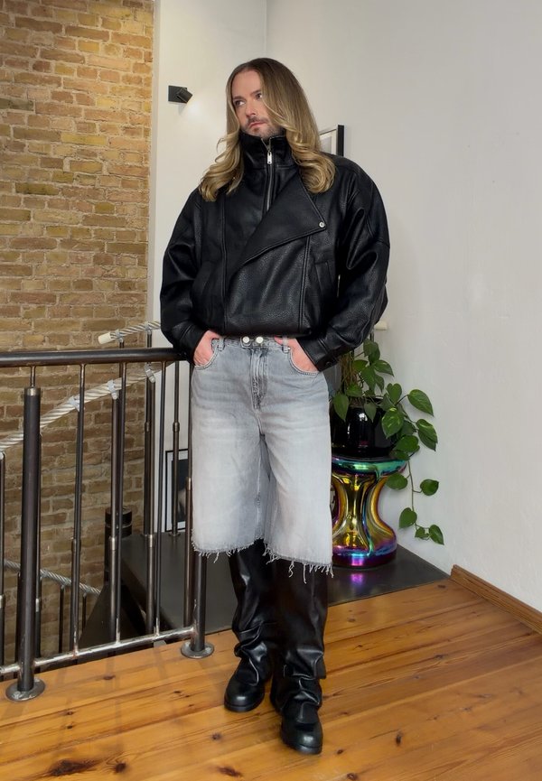 Un hombre de pelo largo posa con las manos en los bolsillos. Lleva una chaqueta de cuero negra, botas y pantalones cortos vaqueros claros con los bajos cortados.