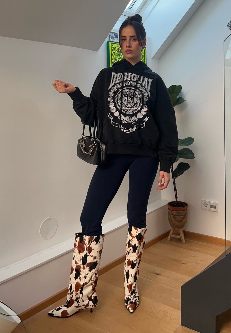 Junge Frau drinnen, trägt einen schwarzen Hoodie, dunkle Hosen, kniehohe Stiefel mit Kuhmuster und hält eine kleine schwarze Handtasche mit dekorativen Perlen.