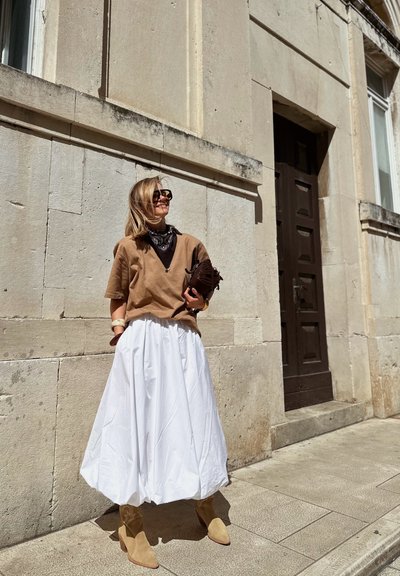 Top marrón de manga corta, falda blanca con tablas, botas de tacón color crema, bandana negra y bolso de mano. De pie contra una pared de piedra texturizada.