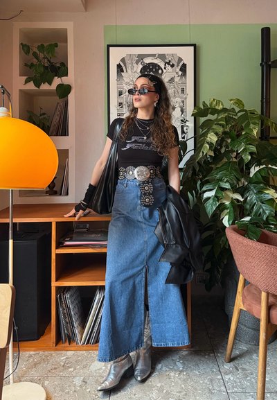 Camiseta negra con letras, falda larga de mezclilla, cinturón plateado con diseños circulares, botas de tobillo plateadas, chaqueta de cuero negra, gafas de sol.