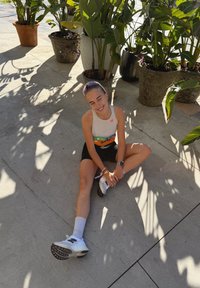 Haut de sport blanc, short noir et baskets blanches avec semelles noires. Assis sur un sol en béton, entouré de plantes vertes en pots.