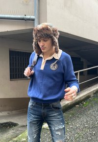 Niebieska bluza z beżowym kołnierzem i okrągłym logo, w zestawieniu z postrzępionymi niebieskimi dżinsami. Na głowie futrzana czapka.