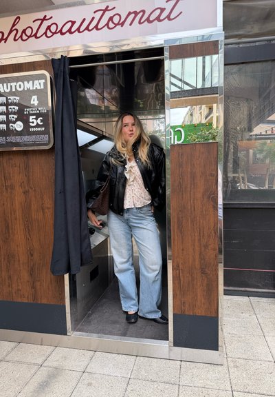 Joven mujer con cabello rubio está de pie en una cabina fotográfica con una chaqueta negra, blusa blanca, vaqueros azul claro y zapatos negros.
