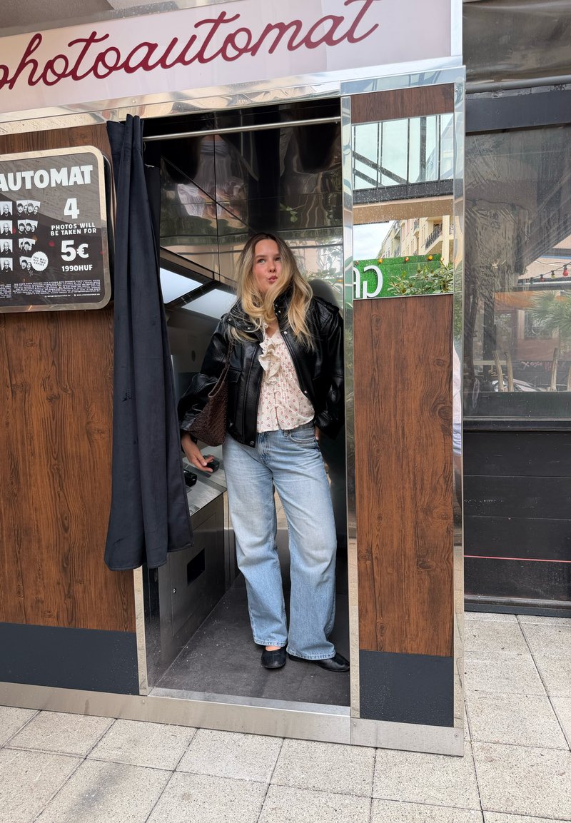 Νεαρή γυναίκα με ξανθά μαλλιά στέκεται σε φωτογραφικό θάλαμο φοράει μαύρο μπουφάν, λευκό πουκάμισο, ανοιχτό μπλε τζιν και μαύρα παπούτσια.