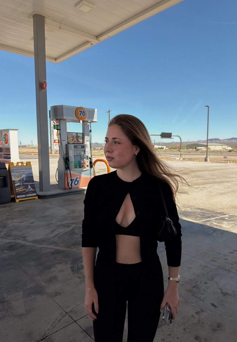 Jeune femme aux longs cheveux se tient dans une station-service 76 sous un auvent, tenant un téléphone, regardant sur le côté par une journée claire et ensoleillée.