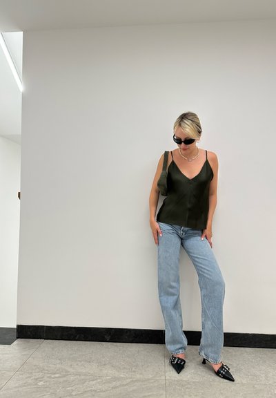 Camiseta de satén verde oscuro con tirantes finos, jeans de pierna ancha de color azul claro y tacones negros de punta con detalles. Fondo neutro.