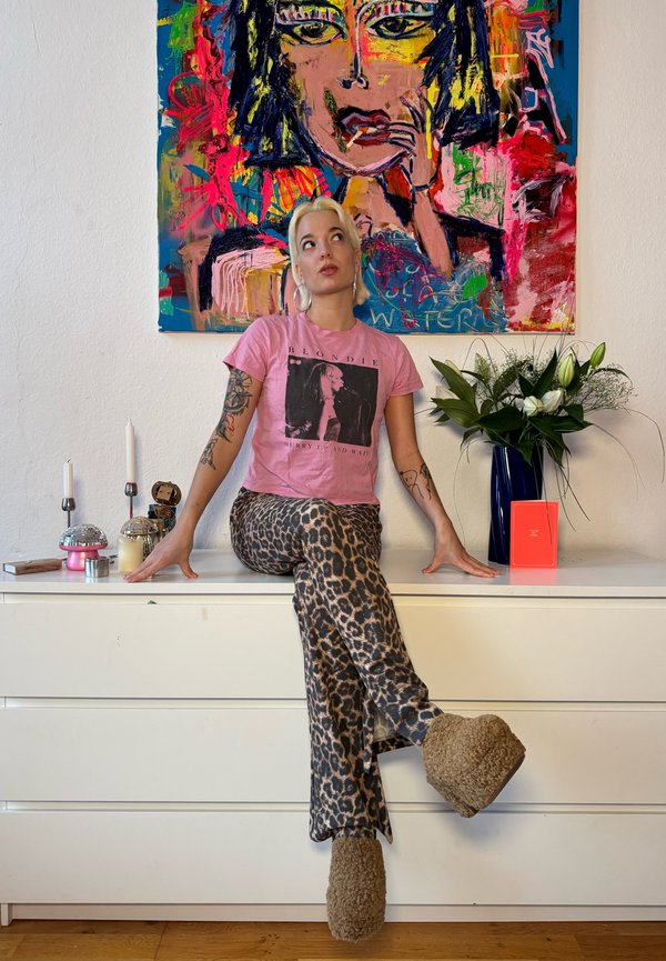 Camiseta rosa con un gráfico negro y pantalones de campana con estampado animal. Zapatitos de plataforma marrones y esponjosos. Pintura abstracta y elementos decorativos en el fondo.