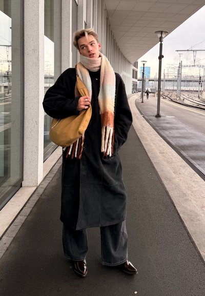 Joven que lleva un abrigo largo negro, bufanda multicolor, lleva un bolso de hombro marrón claro, de pie en el andén de la estación de tren cerca de las vías y el edificio.