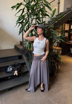 Falda maxi gris con un diseño drapeado, combinada con una blusa sin mangas blanca. Zapatos negros planos y un bolso negro con un llavero de peluche. Entorno interior con plantas.