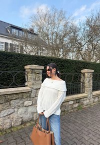 Krēmkrāsas džemperis bez pleciem ar ribotu faktūru, kopā ar gaisā ziliem džinsiem. Tur brūnu rokas somu. Akmens siena un zaļumi fonā.