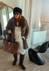 Femme portant un manteau de fourrure sombre, un chapeau noir, des lunettes de soleil, une jupe blanche, des bottes noires, tenant un grand sac à main marron, debout à l'intérieur près d'une chaise noire moderne et d'un miroir.