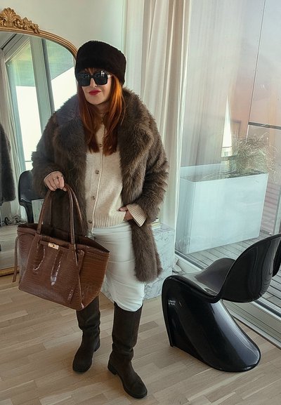 Mujer con abrigo de piel oscuro, sombrero negro, gafas de sol, falda blanca, botas negras, sosteniendo un gran bolso marrón de pie en el interior cerca de una silla negra moderna y un espejo.