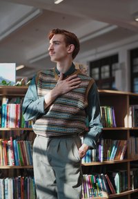 Gilet marrone e blu con motivi sopra una camicia verde, abbinato a pantaloni grigi chiari a pieghe. Un orologio è visibile al polso. Scaffali con libri sullo sfondo.