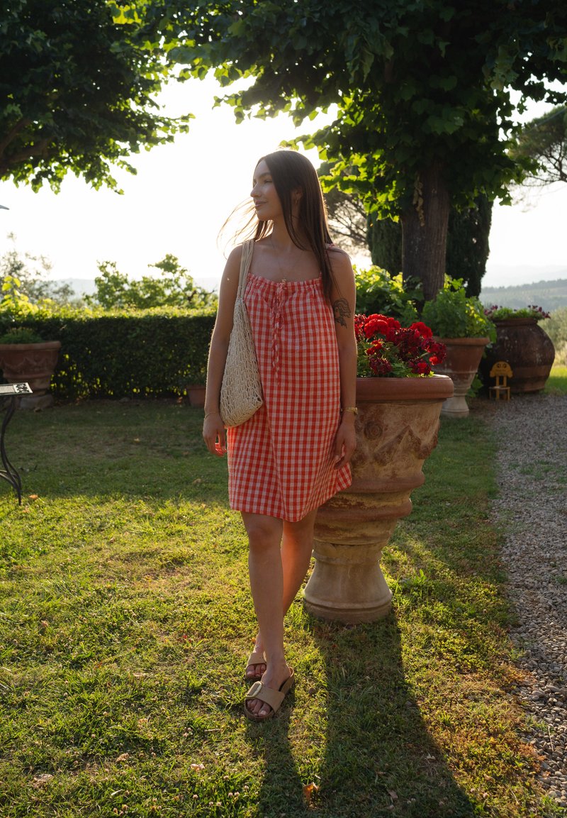 Vestido de gingham vermelho e branco com alças ajustáveis, um corte solto e detalhe de botão, combinado com sandálias castanhas e uma bolsa de palha. Vegetação ao fundo.