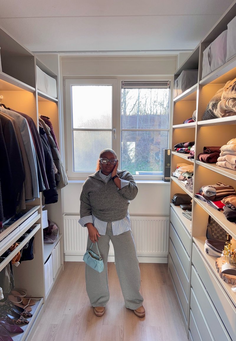 Personne portant un pull gris et un pantalon se tient dans un dressing lumineux, tenant un petit sac bleu, entourée de vêtements pliés et de vestes suspendues.