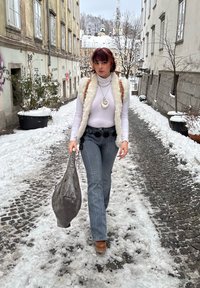 Femme dans une rue enneigée portant un col roulé blanc, un gilet duveteux, un pantalon rayé et un grand sac en cuir gris. Collier remarquable avec un motif en spirale.