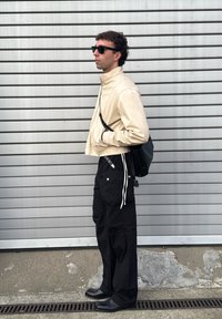 Béžový krátký kabát, černé široké kalhoty s bílými šňůrkami, černá kabelka a černá obuv; modelka má brýle na slunce a stojí před pruhovaným pozadím.