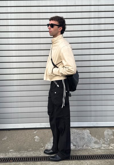 Chaqueta corta beige, pantalones anchos negros con cordones blancos, bolso negro y calzado negro; el modelo lleva gafas de sol y está de pie frente a un fondo a rayas.