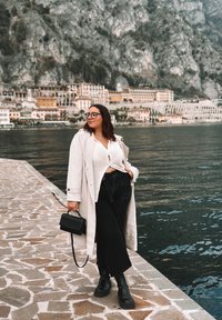 Lungo trench coat beige chiaro, top in maglia bianca, jeans neri a vita alta, stivaletti neri alla caviglia e piccola borsa nera, in piedi vicino all'acqua.