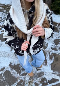 Personne en extérieur portant une veste à motifs noirs et blancs, un jean bleu clair, des chaussures marron clair, ajustant une écharpe blanche en tricot sur un sol enneigé.