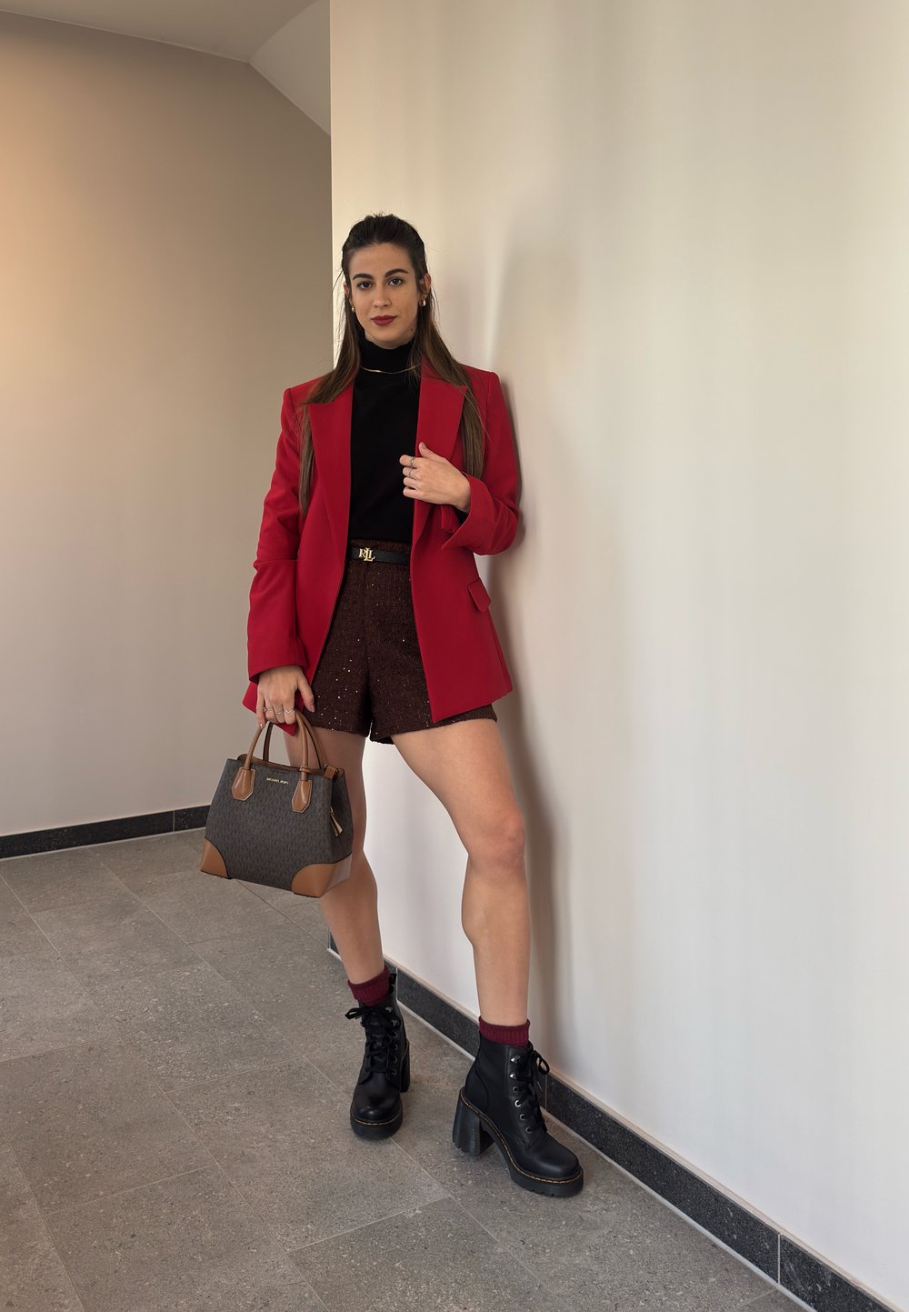 Rød blazer, sort turtleneck, brune strukturerede shorts, sorte snørebåndsstøvletter med hæl og en brun håndtaske med kontrasterende detaljer. Glat væg baggrund.