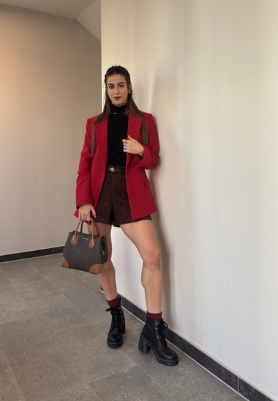 Blazer rojo, cuello de tortuga negro, pantalones cortos texturizados marrones, botas de tacón negras con cordones y un bolso marrón con acentos contrastantes. Fondo de pared lisa.