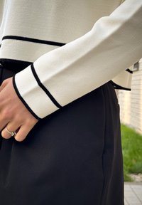 Biely textúrovaný top s čiernym lemovaním na rukávoch, skombinovaný s čiernymi nohavicami. Významné detaily zahŕňajú rebrované manžety a čistý šev.
