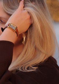 Καφέ δερμάτινο ρολόι με ορθογώνια χρυσή θήκη και μπλε καντράν. Τα μαλλιά πέφτουν γύρω από τον καρπό, αποκαλύπτοντας τα χαρακτηριστικά του ρολογιού.