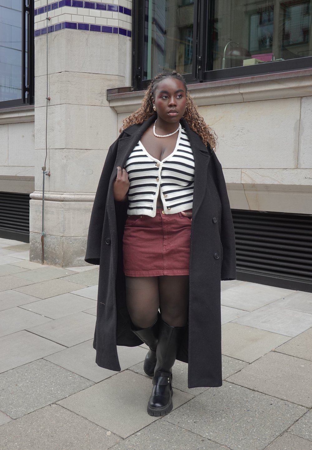 Zwarte overjas, wit en marineblauw gestreepte cardigan, bordeauxrode rok, zwarte panty, hoge schoenen tot de knie. Straatscène, moderne architectuur op de achtergrond.