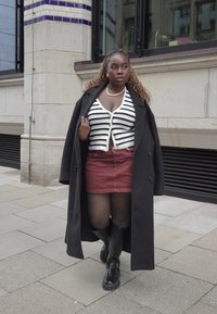 Zwarte overjas, wit en marineblauw gestreepte cardigan, bordeauxrode rok, zwarte panty, hoge schoenen tot de knie. Straatscène, moderne architectuur op de achtergrond.