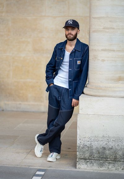 Chaqueta de mezclilla con un bolsillo y costuras blancas, combinada con pantalones oscuros y zapatillas blancas. La figura está apoyada contra una columna.