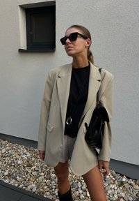 Bež oversized blazer preko črnega majice, kombiniran z črnimi sončnimi očali in črno torbico. Dolg obesek, teksturirana kamnita podlaga.
