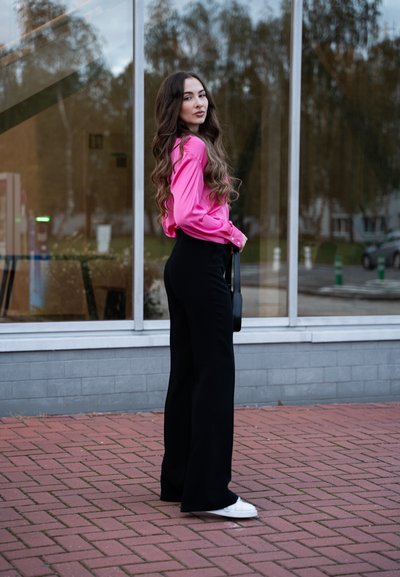 Blusa de satén rosa con mangas largas, combinada con pantalones negros de cintura alta. Zapatillas blancas completan el conjunto. El fondo presenta un edificio de cristal.