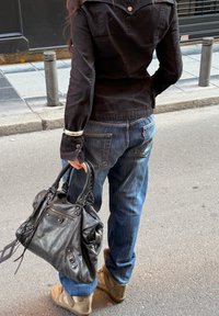 Personne portant une veste foncée et un jean bleu, tenant un grand sac à main en cuir noir, debout sur un trottoir en ville.