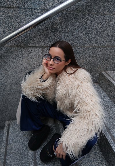 Abrigo peludo de color crema con hebras largas, pantalones deportivos azul marino y zapatos negros. Gafas enmarcan la cara, frente a un fondo de granito.