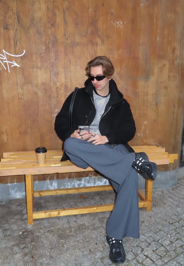 Veste noire ample sur une chemise camouflage grise, pantalon gris à jambes larges, baskets noires avec des bandes blanches, assis sur un banc en bois, tenant un téléphone.