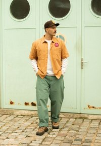 Tan verticaal gestreepte kortemouwen shirt, witte langemouwen shirt eronder, groene cargobroek en schoenen met luipaardprint, staand tegen een mintgroene deur.