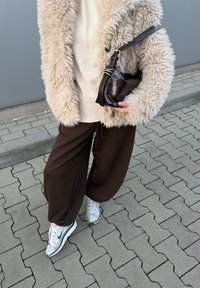 Soft beige coat, cream jumper, brown wide-leg trousers, and silver trainers. Dark brown bag with a short handle held in hand. Background of paved surface.